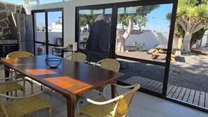An outdoor dining area with a wooden table and wicker chairs, large windows, and a view of a landscaped garden. - Paradise Homes Lanzarote (Nazaret)
