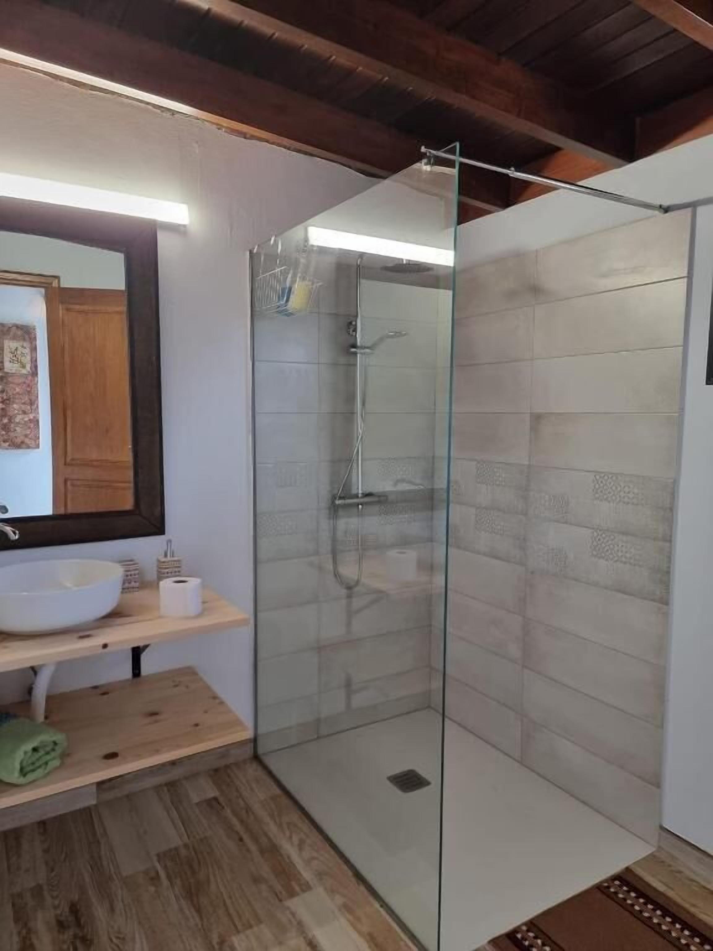 Un baño moderno con ducha envuelta en vidrio, un lavabo blanco y un estante de madera.