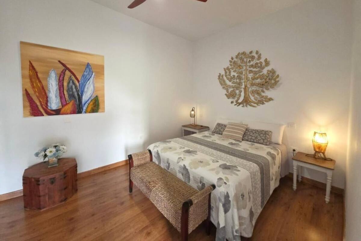 Une chambre à coucher avec un lit, un ventilateur de plafond, une décoration murale en forme d’arbre, une table de chevet avec une lampe et un banc.