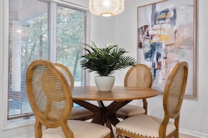 Un'area pranzo con un tavolo rotondo in legno, quattro sedie in rattan, una pianta in vaso e un grande quadro astratto appeso al muro.