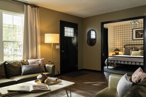 A well-lit room with a sofa, a table with a book and a vase, a lamp, a bed with a headboard, and a door with a window.