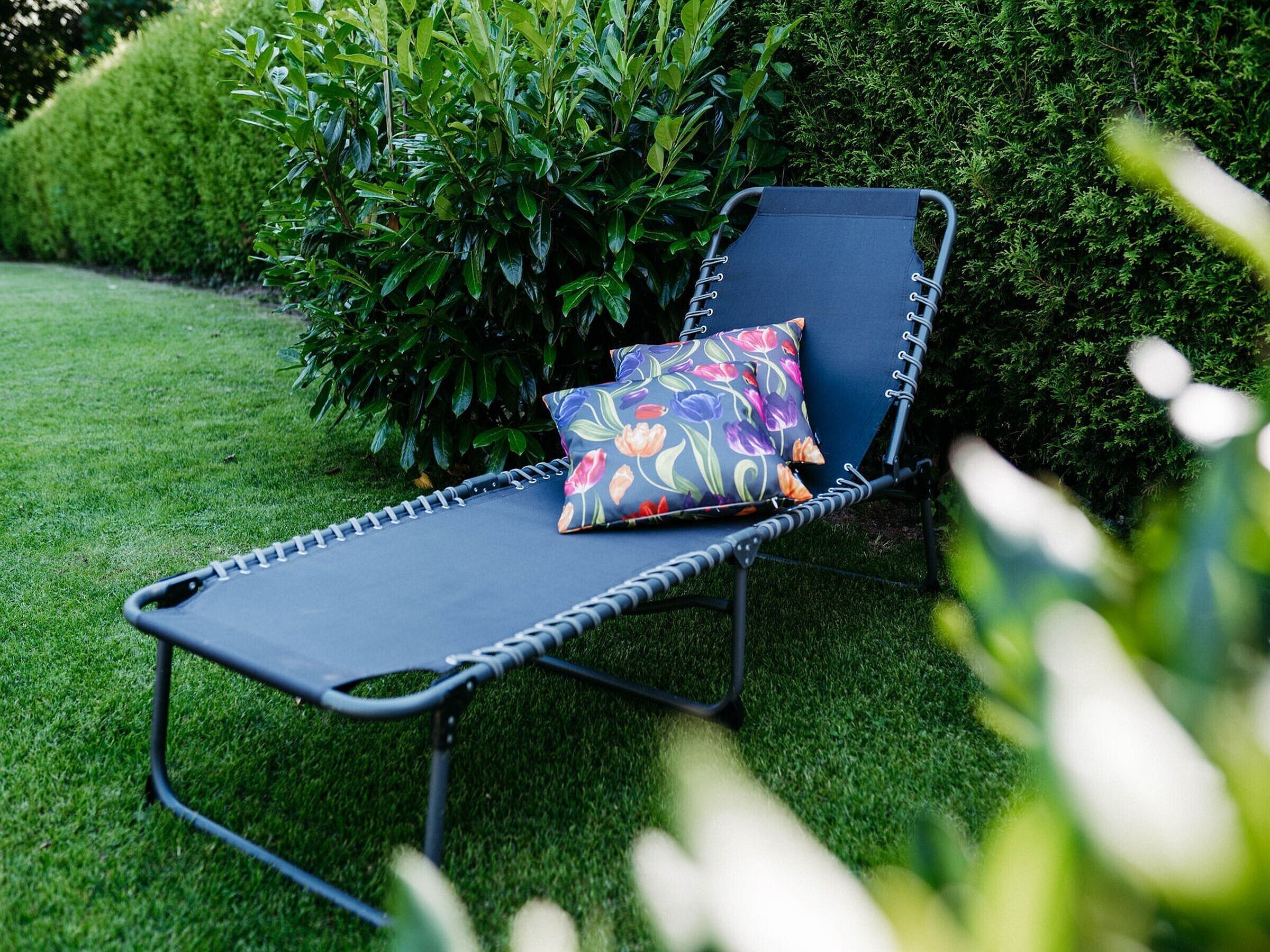 Ein Liegestuhl mit Kissen, platziert in einem Garten mit Gras und Büschen.