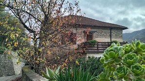 Una casa de piedra con techo de tejas, rodeada de vegetación y un muro de piedra.