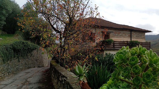 Una casa de piedra con techo de tejas, rodeada de abundante vegetación y colinas.