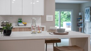 Une cuisine moderne avec des meubles blancs, un îlot central agrémenté d’un vase et de verres, et une vue sur l’extérieur.