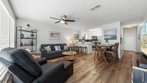 A modern living room with a black leather sofa, a wooden coffee table, and a dining area with a wooden table and chairs. There is a kitchen with white cabinets and a large island. - Gulf Walk by Avantstay West End Coastal Escape Renovated 1BR Condo Shared Pool & Beach Access (Panama City Beach)