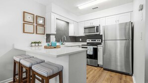 A modern kitchen with white cabinetry, stainless steel appliances, a central island with stools, and decorative wall art. - Gulf Walk by Avantstay West End Coastal Escape Renovated 1BR Condo Shared Pool & Beach Access (Panama City Beach)