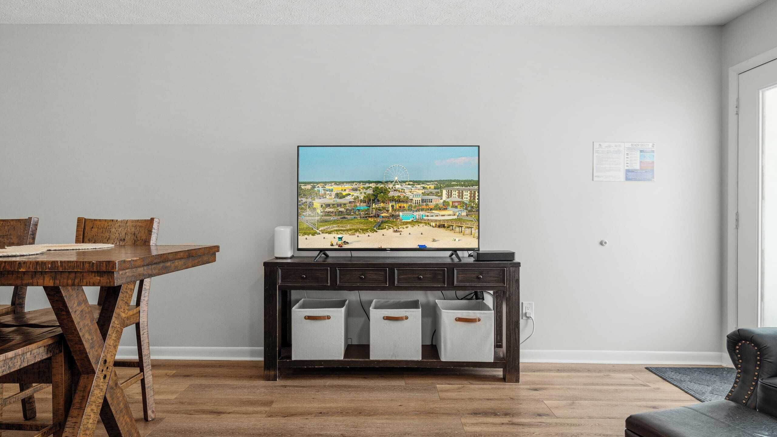Ett vardagsrum med ett matbord i trä, en tv-apparat på ett mörkt träbord och utsikt över en strand med byggnader och ett pariserhjul.