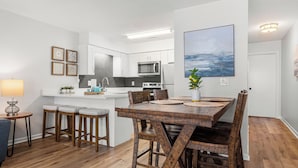 A modern kitchen with white cabinets, a microwave, and a dining area with a wooden table and chairs. - Gulf Walk by Avantstay West End Coastal Escape Renovated 1BR Condo Shared Pool & Beach Access (Panama City Beach)