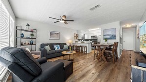 A modern living room with a black leather sofa, a wooden coffee table, and a dining area with a wooden table and chairs. There is a kitchen with white cabinets and a large island. - Gulf Walk by Avantstay West End Coastal Escape Renovated 1BR Condo Shared Pool & Beach Access (Panama City Beach)