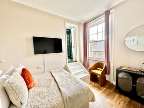 A bedroom with a bed, a TV mounted on the wall, a chair, a small table, and a window with curtains. - Chic studio flat in Camden (Greater London)