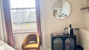 A room with a wooden chair, a round mirror, a small cabinet with a coffee maker, and a window with curtains.
