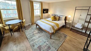 A hotel room with a bed, a round table, a chair, a standing lamp, and a view of buildings outside the window. - Cozy studio Flat in Camden (Greater London)