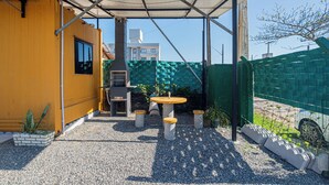 A covered outdoor area with a table and chairs, a barbecue grill, and a view of a building in the background. - Container 3 (Navegantes)