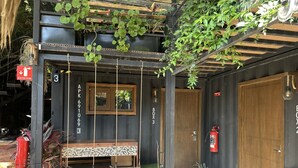 A cozy outdoor seating area with a wooden bench and hanging plants, a fire extinguisher, and a wooden door labeled "BOX 3". - LA SELVA HOSTEL (Mexico City)