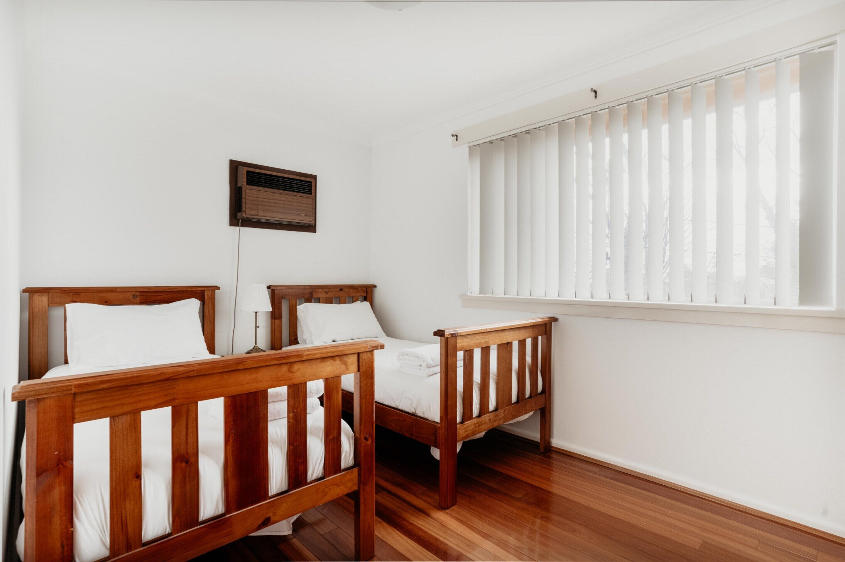 A room with two wooden beds, white walls, a window with blinds, and a wall-mounted air vent.
