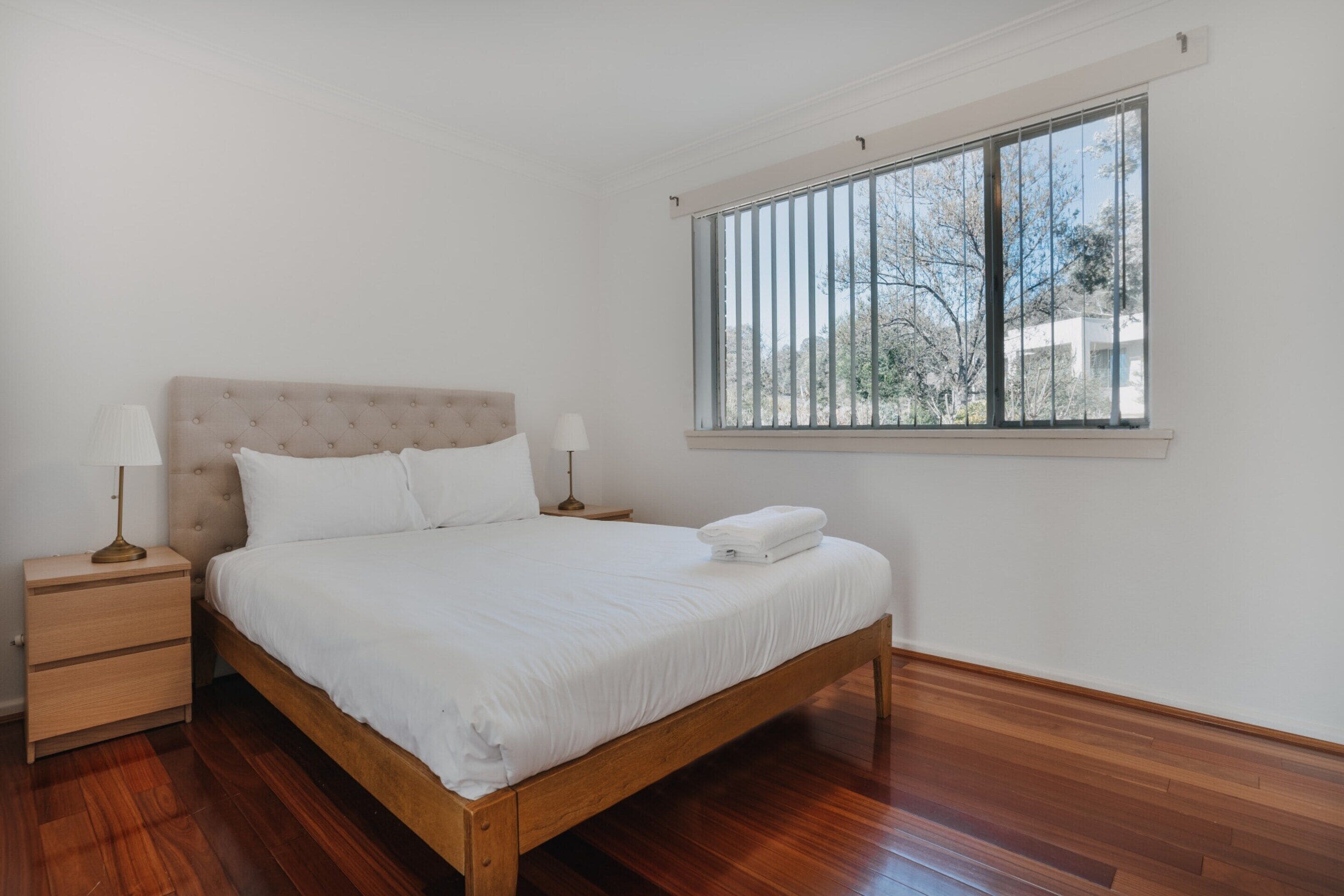 Een slaapkamer met een houten bed, wit beddengoed, een nachtkastje met een lamp en een raam met uitzicht op bomen.