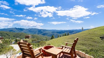A patio with wooden furniture and a brick fire pit overlooking a mountainous landscape.