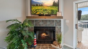 A living room with a fireplace, a television mounted above it, and a view of a balcony with a railing. - Lakeview Condo, Pools, Patio With Grill, Near PB2 Launch (Osage Beach)