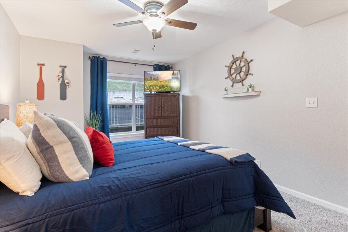 A bedroom with a bed, a ceiling fan, a TV, a ship's wheel decoration, and a view of the outside.