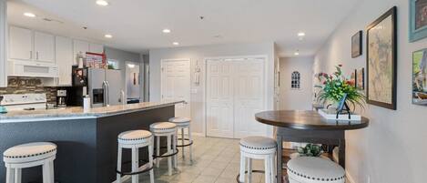 A modern kitchen with white cabinets, a black island, and bar stools. There is a dining table with a plant on it.