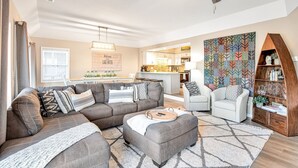 A modern living room with a grey sectional sofa, a matching ottoman, and a wooden side table. There is a patterned rug on the floor, a wooden bookshelf, and a decorative wall hanging. - Captivating Views, Amazing Design and Lake Living (Lake Ozark)