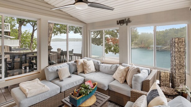 A sunroom with a large window overlooking a lake, a ceiling fan, and a cozy seating area with cushions and a woven coffee table. - Captivating Views, Amazing Design and Lake Living (Lake Ozark)