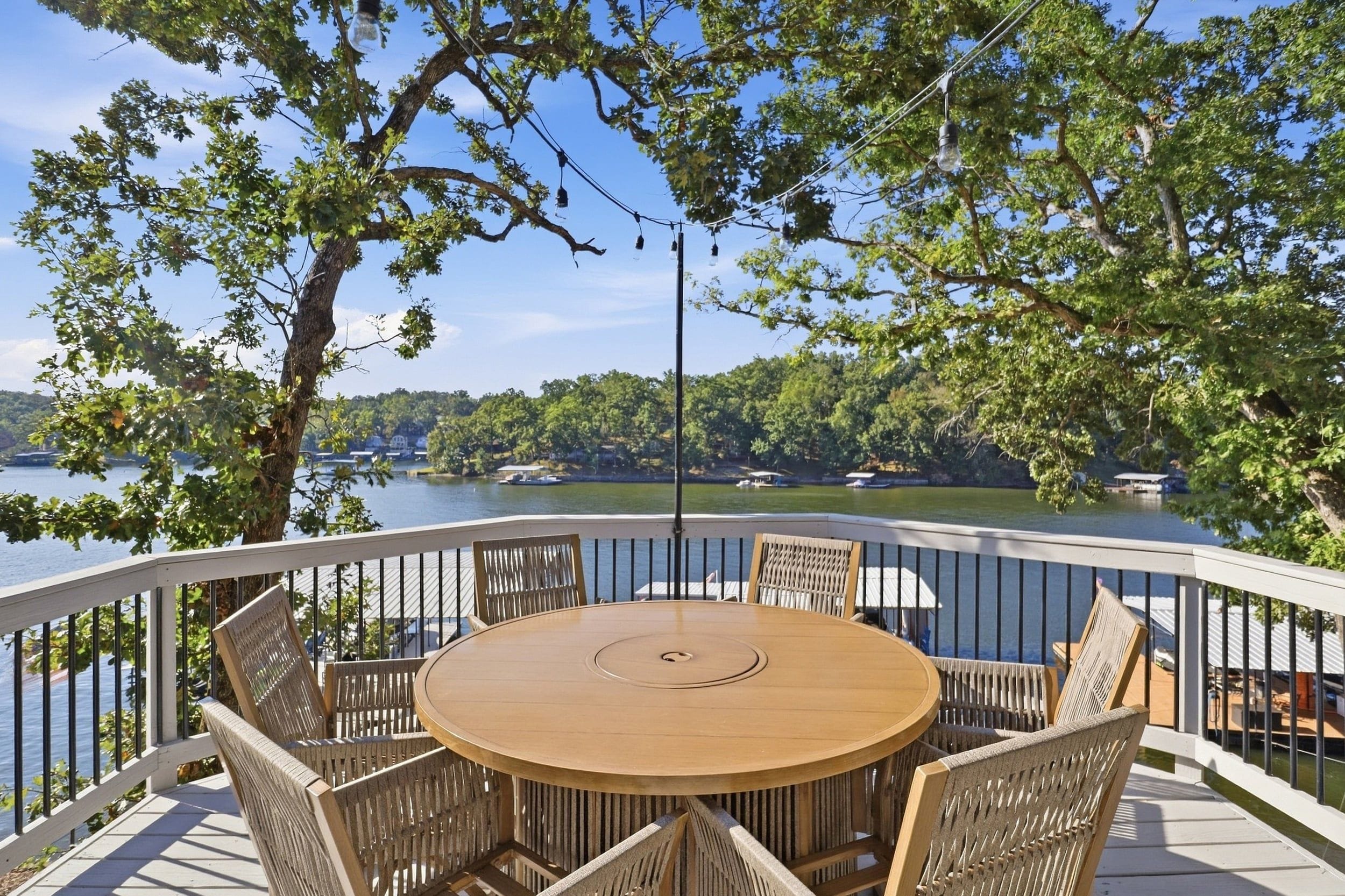 A round wooden table with six chairs on a balcony with a white railing, overlooking a lake with boats and a forested shoreline.