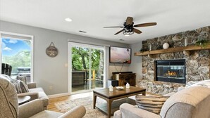 A living room with a stone fireplace, a ceiling fan, and a large TV.