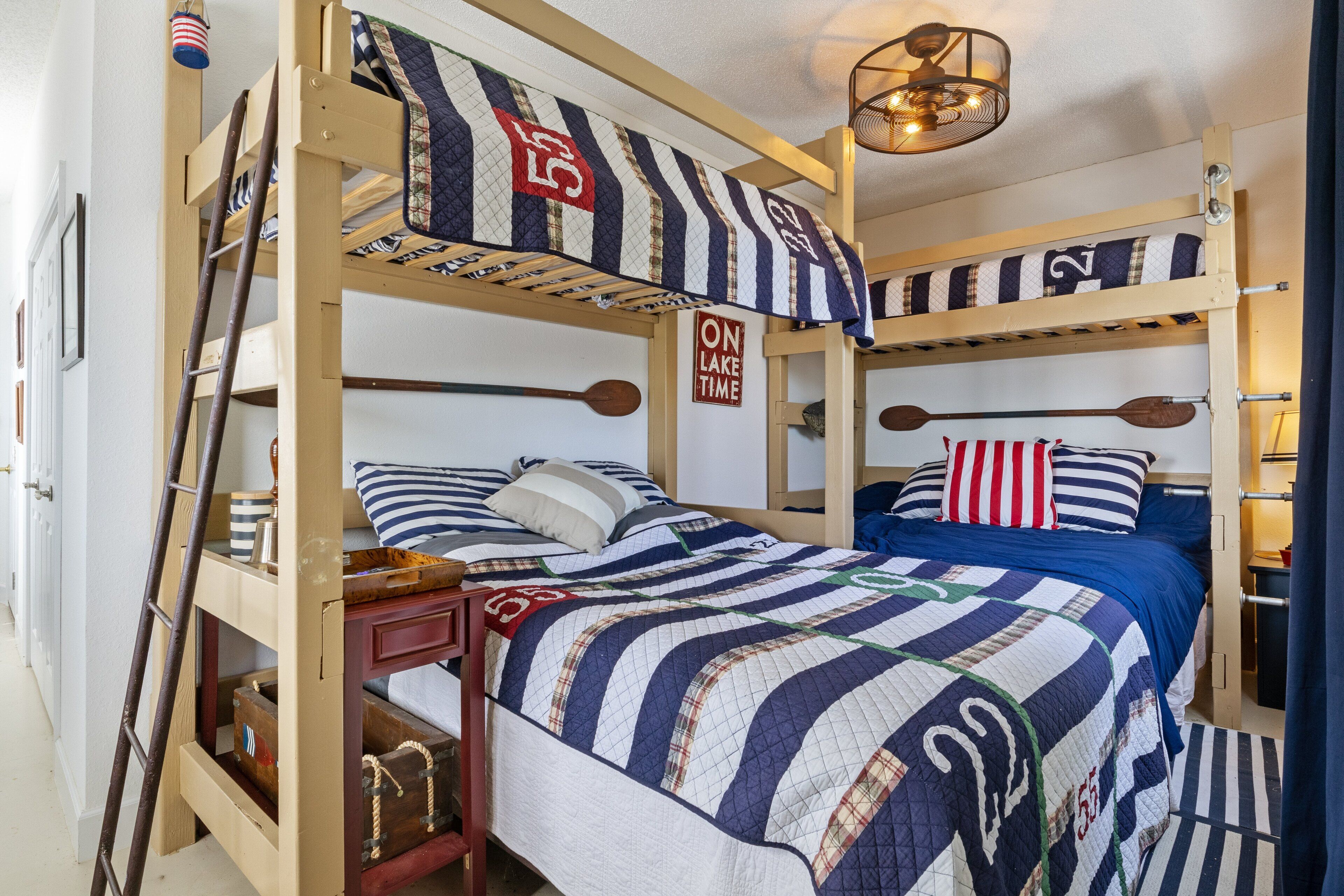 Une chambre avec un lit superposé, une literie à rayures, une échelle, un panneau indiquant « ON EST EN MODE VACANCES AU LAC » et une tête de lit en bois.
