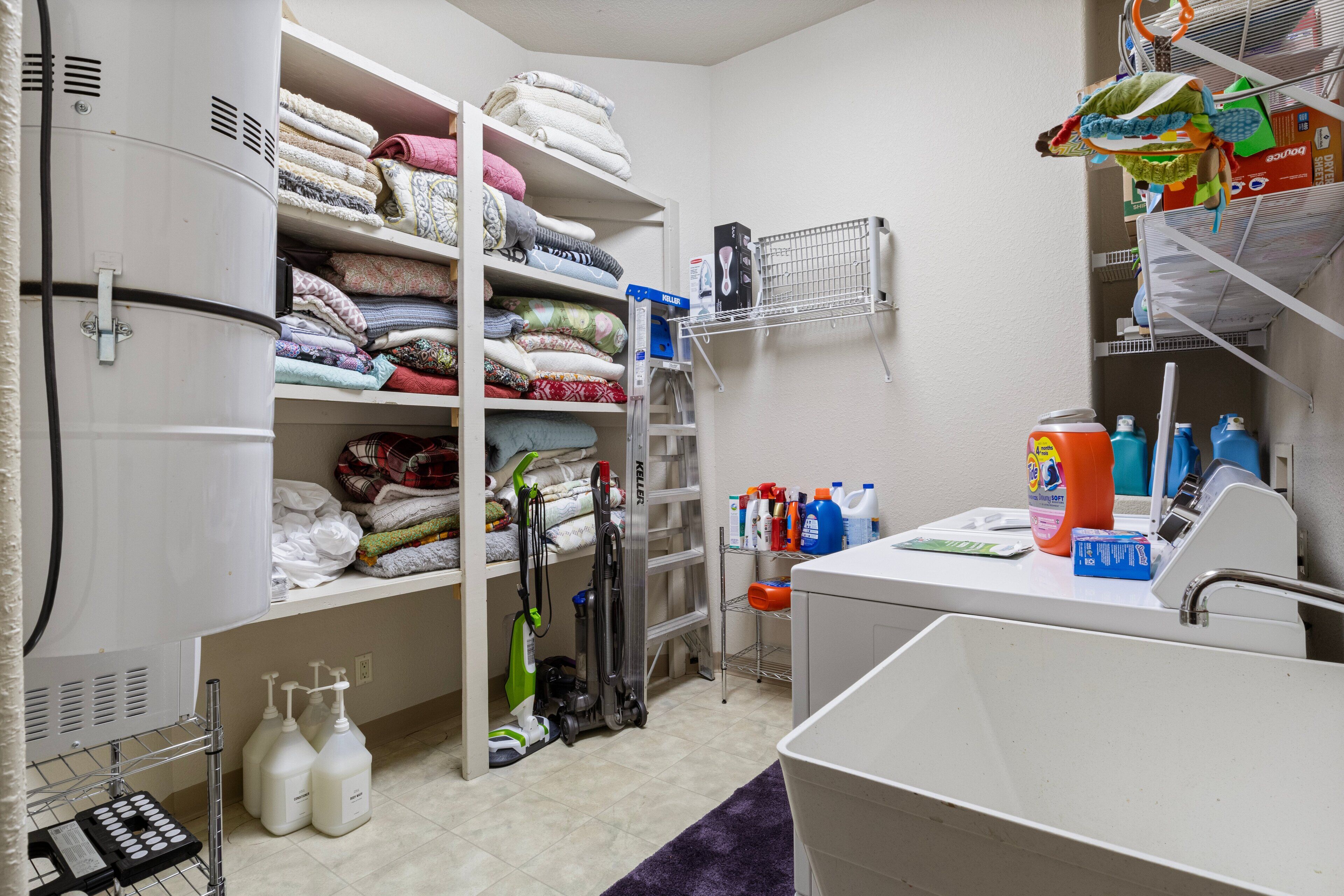 A laundry room with shelves of folded towels, a washing machine, a dryer, and various cleaning supplies.