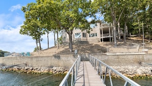 A concrete dock leading to a building with a balcony, surrounded by trees and a rocky shoreline. - Pet-friendly Home In The Main Channel w/ Breathtaking Sunsets & Lake Views (Lake Ozark)