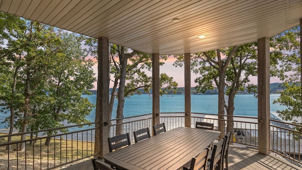 A covered outdoor deck with a wooden table and chairs, overlooking a body of water and trees. - Pet-friendly Home In The Main Channel w/ Breathtaking Sunsets & Lake Views (Lake Ozark)