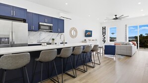 A modern kitchen with dark blue cabinets, a white countertop, and a built-in stove. There are four bar stools and a large window offering a view of the outdoors. - Expansive Deck, Private Dock, Boat Slip and Stunning Views (Climax Springs)