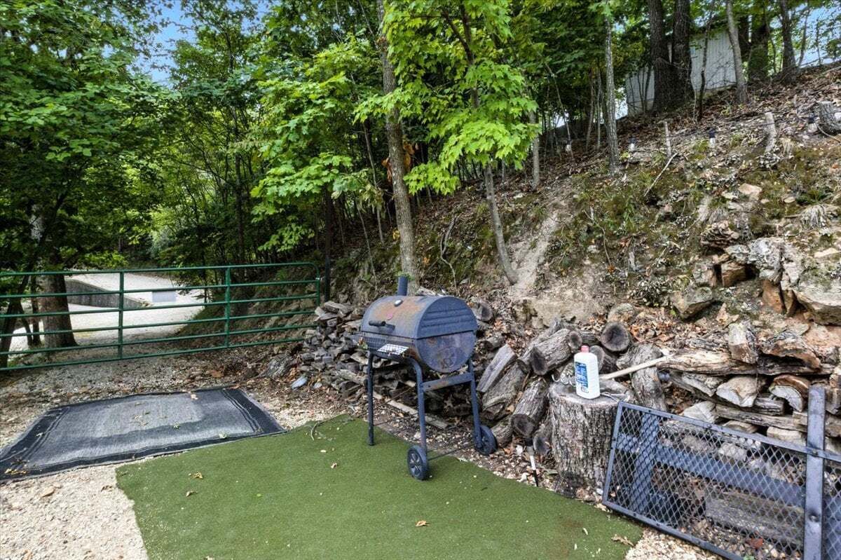 A barbecue grill in a wooded area with a green metal fence, a small patch of artificial turf, and a white bottle on a tree stump.