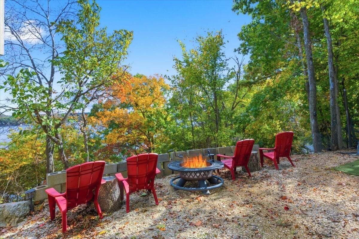 Un espace extérieur aménagé avec des sièges, un foyer et des chaises rouges, entouré d’arbres et offrant une vue sur une étendue d’eau.