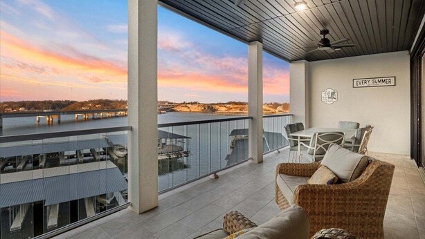 En balkon med udsigt til en flod og solnedgang, fletmøbler og et skilt med teksten "EVERY SUMMER".