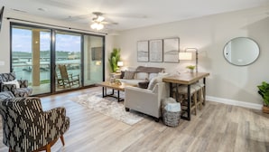 Un salon moderne comprenant un canapé, des fauteuils, une table basse et une table à manger. Une grande fenêtre offre une vue sur un pont et sur la verdure environnante.