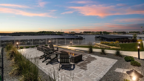 Un espace extérieur aménagé en terrasse, avec des sièges et un foyer, entouré d’un jardin paysager et offrant une vue sur une étendue d’eau.