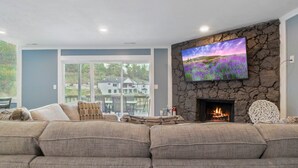 A living room with a stone fireplace, a large TV mounted on the wall, and a view of a house through a glass door. - Our Cove Retreat - Perfect for Wave-free Swimming, Tanning & Prime Location Fun! (Lake Ozark)