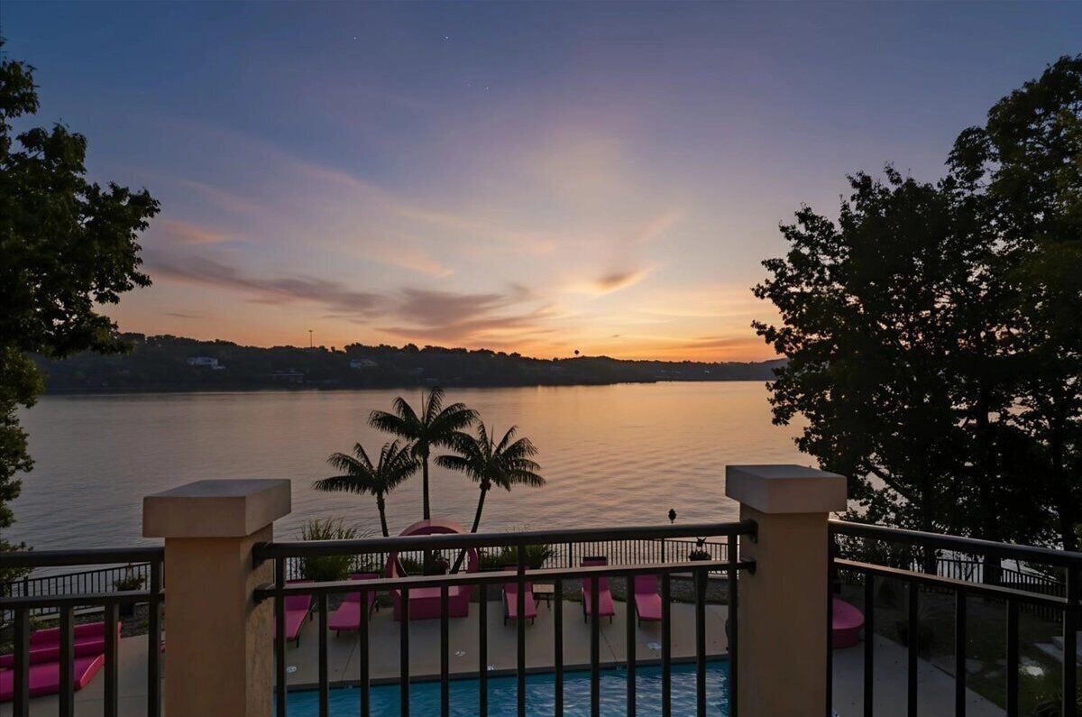 A poolside view with a sunset over a body of water, palm trees, and a distant shoreline.