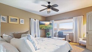 A hotel room with a large bed, a desk, a chair, a TV, and a view of the sea through a window.