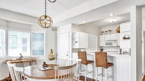 A dining area with a wooden table and white chairs, a kitchen with white cabinets and a microwave, and a hanging light fixture. - Breathtaking Views - Two Pools - Enclosed Balcony (Lake Ozark)