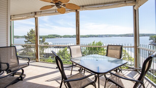 A balcony with a glass table and chairs, overlooking a body of water and a distant bridge. - Breathtaking Views - Two Pools - Enclosed Balcony (Lake Ozark)