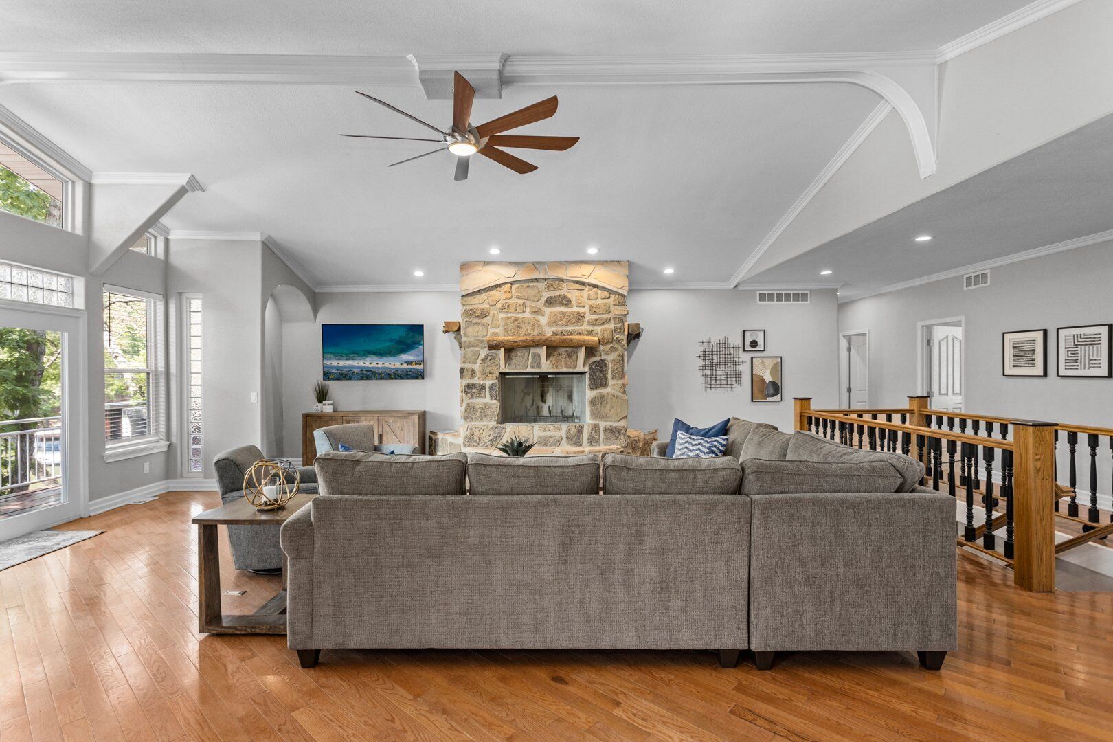 A spacious living room with a stone fireplace, a large gray sectional sofa, and a wooden staircase.
