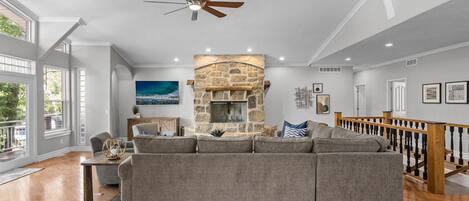 A spacious living room with a stone fireplace, a large gray sectional sofa, and a wooden staircase.