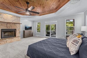 A bedroom with a stone fireplace, a bed with a quilted cover, a ceiling fan, and a view of the outdoors through a glass door.