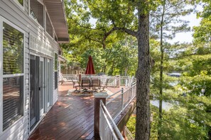A wooden deck with a dining table and chairs, a red umbrella, and a railing overlooking a body of water.