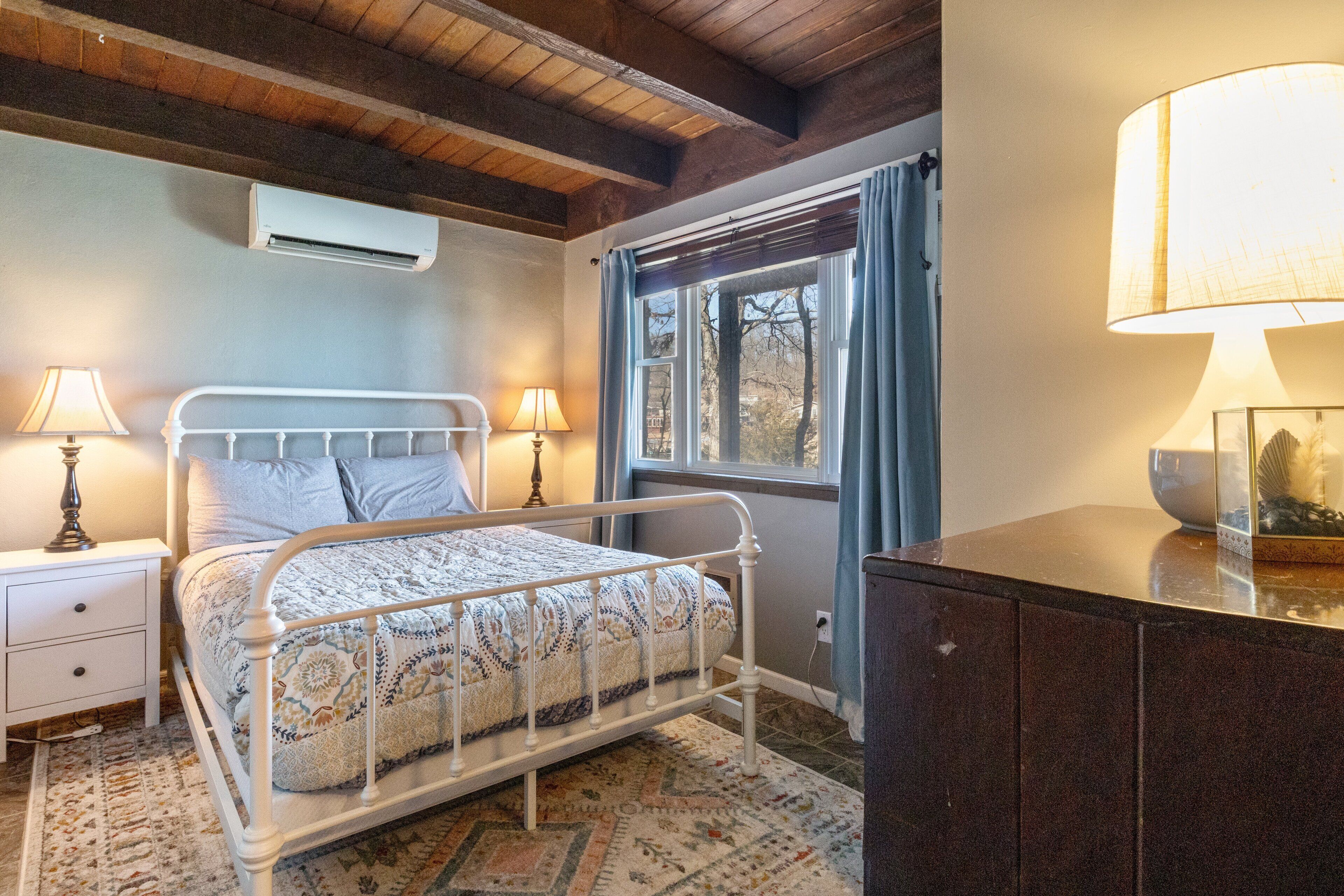 Een slaapkamer met een metalen bed, twee nachtkastjes met lampen, een raam met gordijnen en een houten plafond.