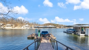 A wooden dock leading to a covered boat launch area with a view of a calm lake and distant buildings. - Fishing, Boat Slip, Morning Coffee, Picturesque View (Sunrise Beach)